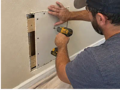 Handyman fastening a supported drywall patch into a wall opening above protected tile flooring