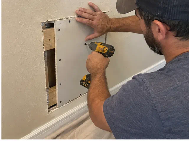 Handyman fastening a supported drywall patch into a wall opening above protected tile flooring